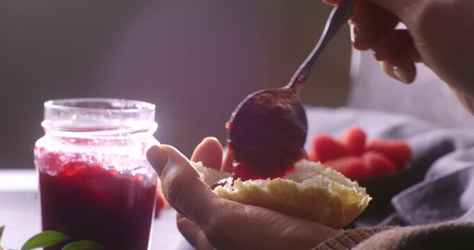A hand carefully spoons rich raspberry jam from a glass jar onto freshly torn bread. Soft lighting emphasizes the vibrant color and smooth texture of the jam, with raspberries in the background.