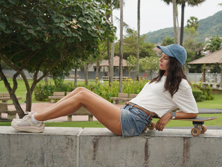 A young woman sits comfortably on a low wall at the park holding a skateboard in her hands