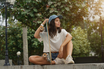 A young woman sits comfortably on a low wall at the park holding a skateboard in her hands