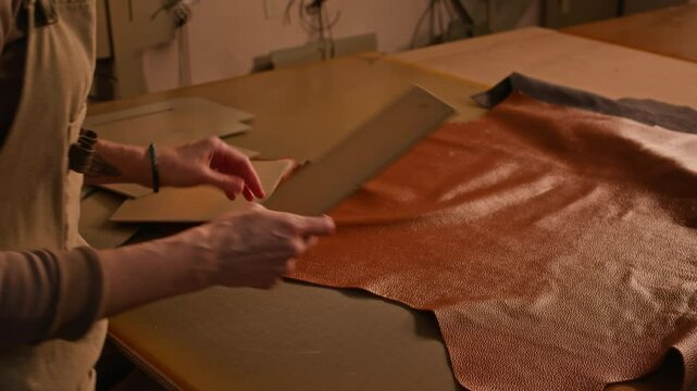 Side view cropped shot of unrecognizable tanner wearing apron choosing pattern for camel leather bag production working in dim lit professional workshop