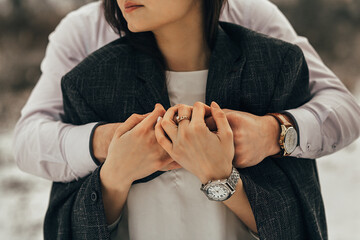two people. A woman stands with her back to a man who hugs her. The woman has long dark hair and is wearing a dark coat and white blouse. She wears rings on her left hand and a silver watch on her rig