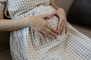 A woman is sitting on a couch with her hands on her stomach, making a heart shape. She is pregnant and she is in a relaxed and comfortable mood
