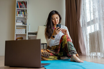 Asian woman in colorful afghani pants sitting on yoga mat with phone