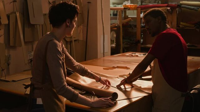 Medium shot of young Caucasian craftswoman teaching young Black male apprentice to cut leather textile using special tanning tools working together in cozy dim lit authentic craft studio