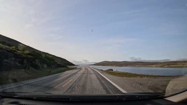 Driving along Norway's E69 road in the north. Rugged rocky cliffs on the right, the sea on the left. A band of clouds and mist hovers above the water, creating a serene and dramatic scene.