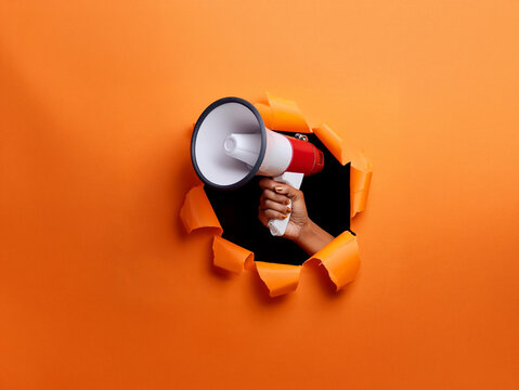 A hand holds a megaphone through a torn orange background, announcing a message.  The vibrant color contrasts with the black hole, creating a striking visual.