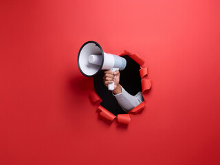 A hand holding a megaphone bursts through a red paper background, announcing an urgent message.  The scene is minimalist and impactful, symbolizing a powerful announcement or call to action.