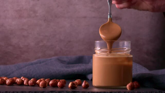 Smooth hazelnut butter drizzles from a spoon into a glass jar. Whole hazelnuts are scattered on a dark surface, with a soft blue fabric in the background adding depth and warmth.