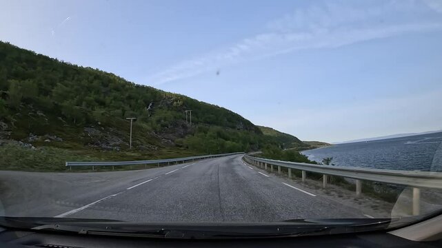Driving through the tundra on E69 in northern Norway. Rocky hills with sparse trees and grass rise on the left, while the sea stretches along the right, showcasing raw and rugged nature.