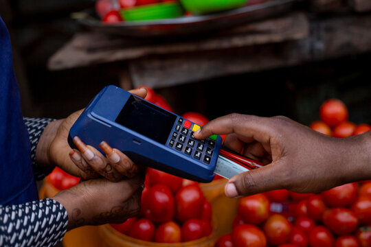 A hand using a bank card on a point-of-sale terminal at a market stall selling fresh tomatoes, representing digital financial transactions and modern commerce in traditional outdoor settings.