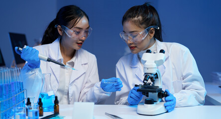 Two female scientists wearing lab coats and safety glasses are conducting research in a modern...