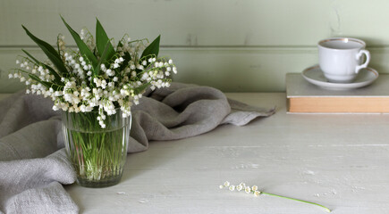 Lily of the valley bouquet in glass vase on white table with teacup and book