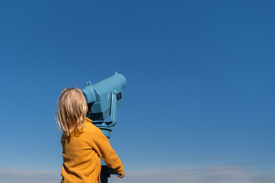 Child looking through a blue telescope under a bright blue sky.