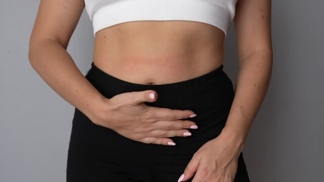 Young woman stroking her belly in a circle with her hand to speed up the digestion of food on grey background. Health care, good digestion. The girl shows a feeling of hunger. Overeating. Slow motion