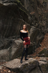 The girl stands with her hands on her hips, while rocky cliffs and a reflection in the water are visible in the background. The landscape around looks like autumn, with bare trees and gray rocks.