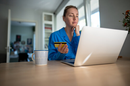 Focused woman on laptop uses credit card for online transactions, lost in heavy spending. Credit debt, online shopping addiction, financial strain, insecure transactions, fraud risk, impulsive buying