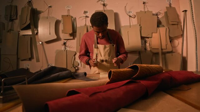 Medium shot of concentrated young African American craftsman at work in authentic tannery among leather rolls and patterns, using punch and hammer for perforation