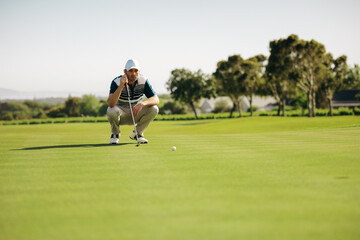 Crouching golfer on putting green aiming at golf ball on a lush green course under a clear sky,...