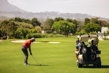 Man aiming for the green near sand bunker, with friend relaxing in golf cart