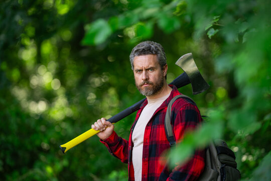Hard working man with axe in forest. A bearded lumberjack with a large ax. Portrait of a brutal bearded man in a plaid shirt holds an axe. Male beauty and masculinity.