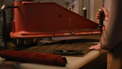 Side view cropped shot of unrecognizable female tanner wearing apron cutting leather details using special press standing at desk in dim lit workshop