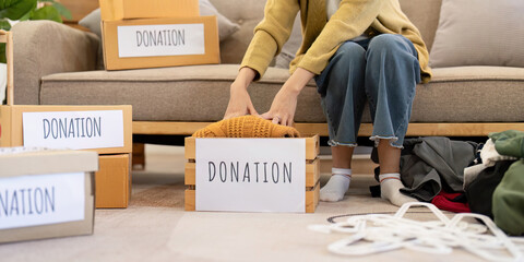 Woman gently placing a knitted sweater into a donation box, organizing clothes in a warm living room