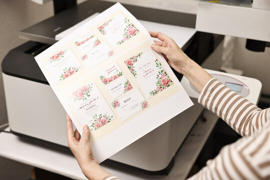 Woman with printed invitation cards near modern printer indoors, closeup. Printing house