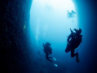 silhouette of unrecognizable scuba divers with flashlights swimming in narrow dark cave with rough rocky formations undersea in deep clear water, concept of active leisure and underwater adventure