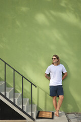 A man casually leaning against a vibrant green wall beside a set of stairs