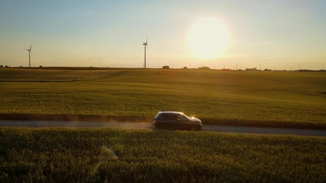 SUV driving along dirt road through green fields, leaving trail of dust behind under warm evening sunlight. Auto cruising through countryside as golden sunlight highlights dust cloud trailing car. Car