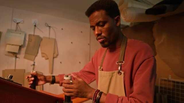 Tilt up shot of concentrated young African American craftsman in apron using leather cutting machine standing at workbench in well-equipped dim lit crafting studio with patterns on wall