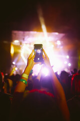 Hand with a phone records live music festival. Audience crowd of people use smart phones enjoying...