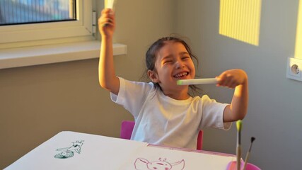 a little child girl happily makes prints in an album with her hands with a dirty face and smiles, a happy child draws a picture with gouache fingers at home at the table. High quality 4k footage