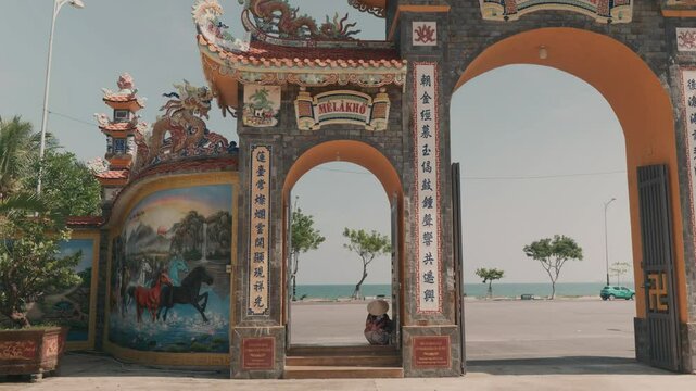 Vietnamese woman resting in traditional conical hat near ornate temple entrance, decorative dragons surrounding, colorful mural featuring horses, turquoise car parked beside coastal road