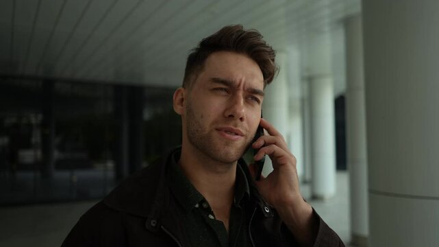 Close-up face of focused male wearing casual attire talking on smartphone, standing on city street with concentrated demeanor, looking away with serious expression. Shooting in slow motion.