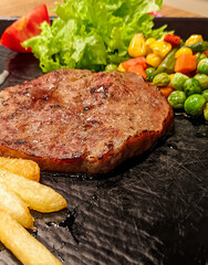 Close up photo of sirloin steak served with vegetables and potatoes on a black platter