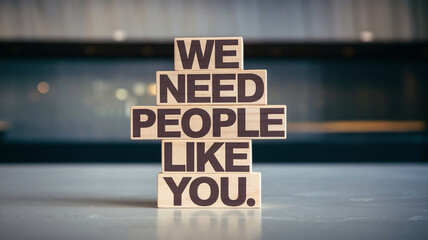 A stack of wooden blocks displays the phrase "WE NEED PEOPLE LIKE YOU" in bold, dark brown letters. Set on a flat surface with a blurred indoor background, it conveys a motivational, recruitment tone.