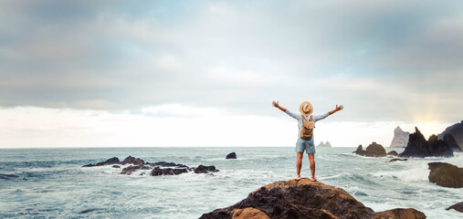 Man with backpack raising arms up at the beach - Traveler male enjoying freedom on summer vacation - Successful hiker celebrating victory on the top of the mountain