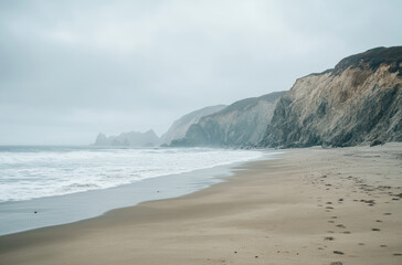 Secluded beach stretches alongside steep cliffs while gentle waves lap at the shore. A gray, cloudy sky looms above, adding a serene and tranquil atmosphere to the coastal scenery