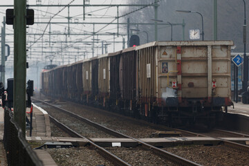 Train passing through a busy station with cargo wagons and commuters on a misty morning