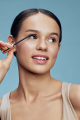 Young woman applying mascara with a smile against a light blue background, showcasing natural beauty and makeup techniques for a fresh look