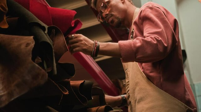 Low angle shot of experienced young African American artisan selecting leather among multiple fabric rolls of different colors and sizes storing on shelves in tannery