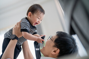 Father joyfully lifts his laughing baby boy in a bright indoor setting on a cheerful afternoon