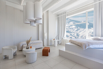 modern pure white interior design of a spacious room features a cozy bed and stylish furniture. Large windows frame a stunning snowy mountain landscape, creating a serene atmosphere during daylight.
