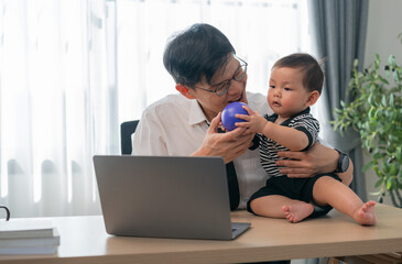 Grandfather plays with baby at home office while working at a laptop on desk