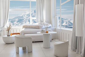 modern pure white interior design of a spacious room features a cozy bed and stylish furniture. Large windows frame a stunning snowy mountain landscape, creating a serene atmosphere during daylight.