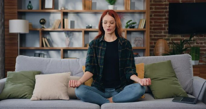 Woman doing yoga in lotus position and closing eyes of meditation sits on sofa at home. Practice feels inner harmony and balance no stress.
