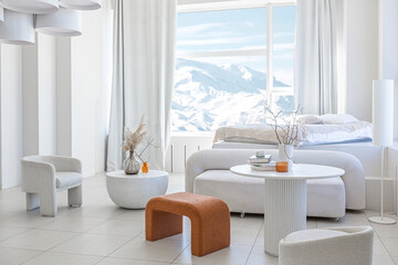 modern pure white interior design of a spacious room features a cozy bed and stylish furniture. Large windows frame a stunning snowy mountain landscape, creating a serene atmosphere during daylight.