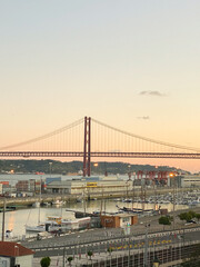 Fototapeta premium Sunset view of the iconic red suspension bridge over the river, marina with yachts, industrial port, and clear sky.