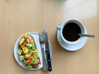 Top view of avocado toast with cherry tomatoes on a plate, served with a cup of black coffee on a wooden table.
